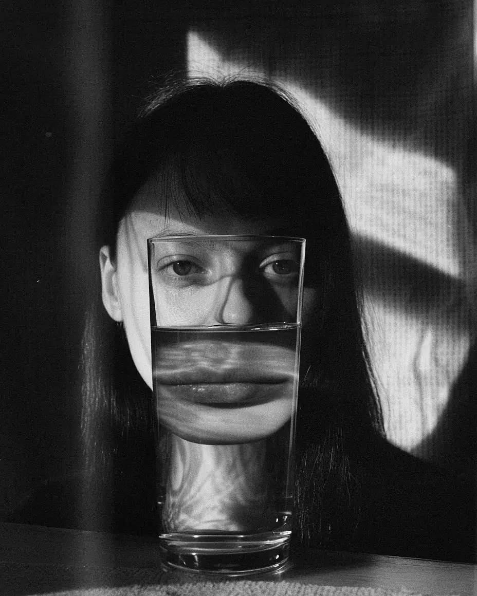 Retrato em preto e branco de uma mulher atrás de um copo d'água, com luz dura lateral projetando o desenho de uma janela na parede